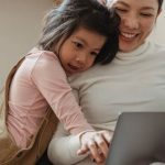 User-generated Content - Cheerful smiling Asian woman browsing modern netbook while hugging with cute content daughter on comfy sofa in cozy living room