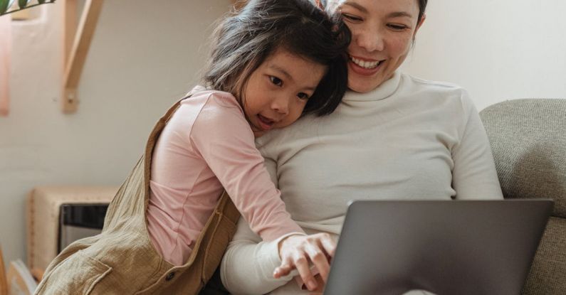 User-generated Content - Cheerful smiling Asian woman browsing modern netbook while hugging with cute content daughter on comfy sofa in cozy living room