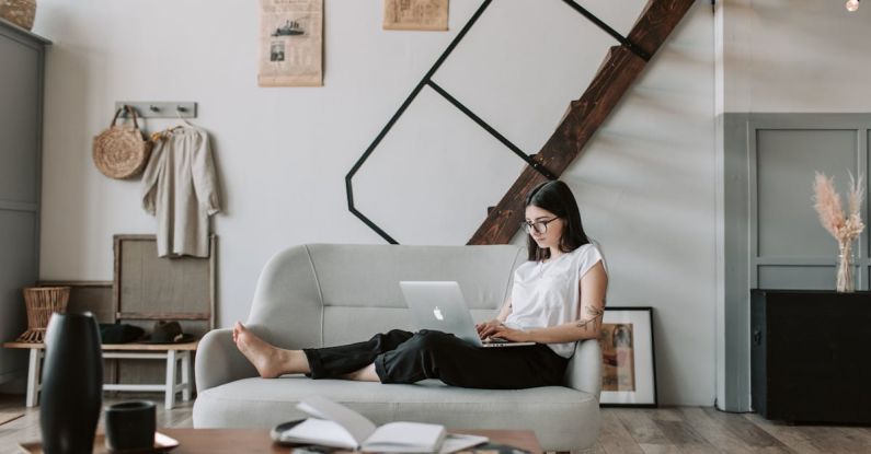 Ecommerce Email - Positive female with tattooed arms wearing casual clothes and eyeglasses reading netbook while sitting barefoot on comfortable sofa behind table with books and magazines in modern apartment with minimalist interior in eco style during free time