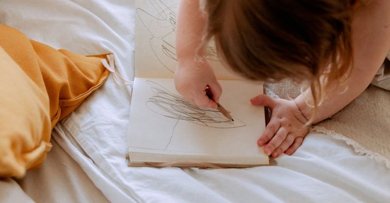 Ecommerce Small Business - From above of small girl in dress drawing with pencil in notebook while mother using laptop on bed near