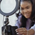 Content Influencer - Cheerful young African American female blogger in stylish sweater smiling while setting up camera of smartphone attached to tripod with ring light before recording vlog