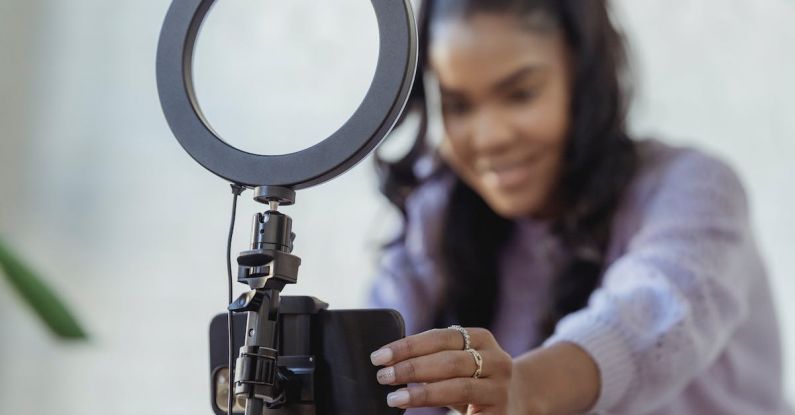 Content Influencer - Cheerful young African American female blogger in stylish sweater smiling while setting up camera of smartphone attached to tripod with ring light before recording vlog