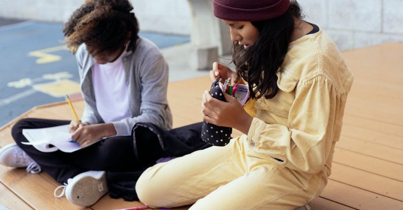 Case Study Influencer - Crop Hispanic schoolgirl with pencil case sitting near anonymous black girlfriend writing in copybook while doing homework on wooden platform