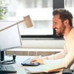 Email Metrics - Man in White Dress Shirt Sitting on Black Rolling Chair While Facing Black Computer Set and Smiling