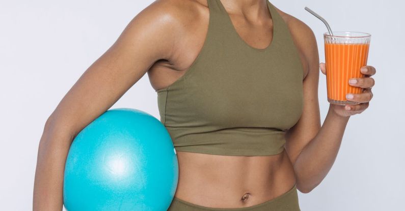 Content Recycling - Cheerful African American lady in activewear with blue ball drinking orange juice and looking at camera against white background